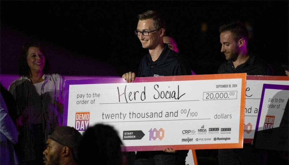 A group of people smile and celebrate as two men hold a large presentation check made out to "Herd Social" for \$20,000. The check is from Demo Day 2024, with logos from sponsors like Meijer, PNC Bank, and Huntington visible at the bottom.